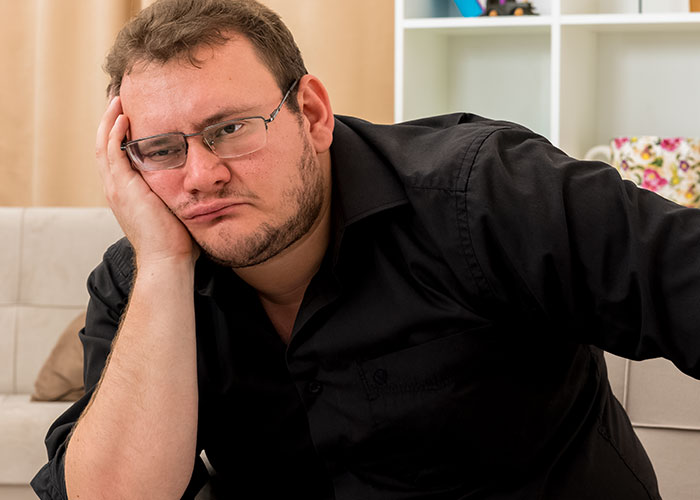 Man sitting on a couch, wearing glasses and a black shirt, looking tired and contemplative indoors.