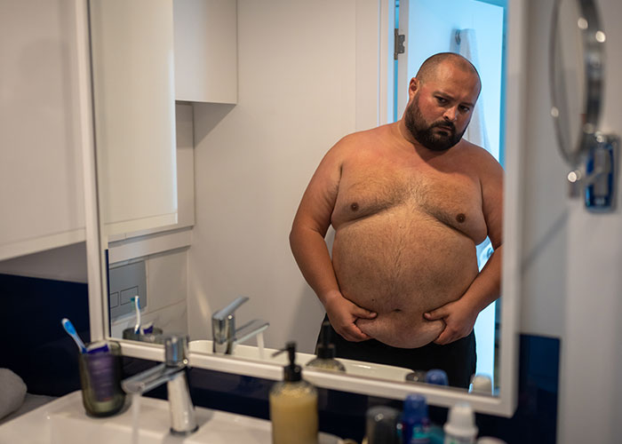 Overweight man looking at himself in the bathroom mirror, contemplating.