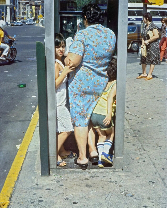 Street photography: Woman and children in a phone booth on a busy city sidewalk.