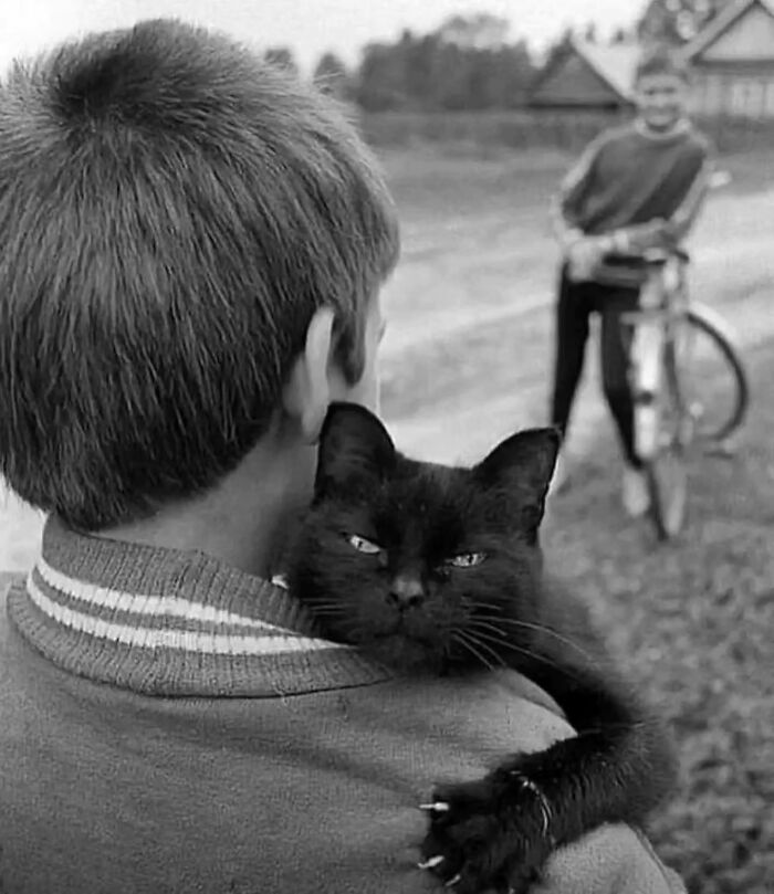 Boy holding a black cat on his shoulder in a street scene, with a cyclist in the background.