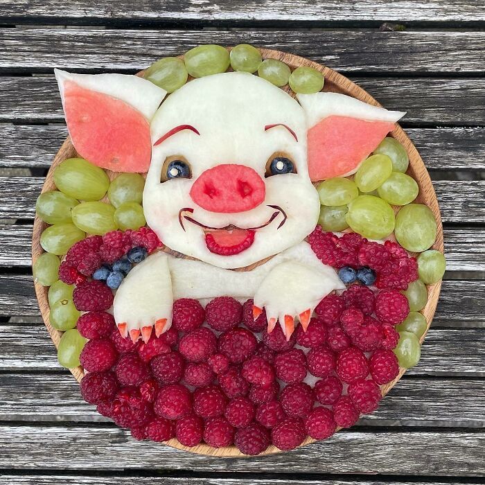 Healthy eating art featuring a pig made from fruit, surrounded by grapes, raspberries, and blueberries on a wooden table.