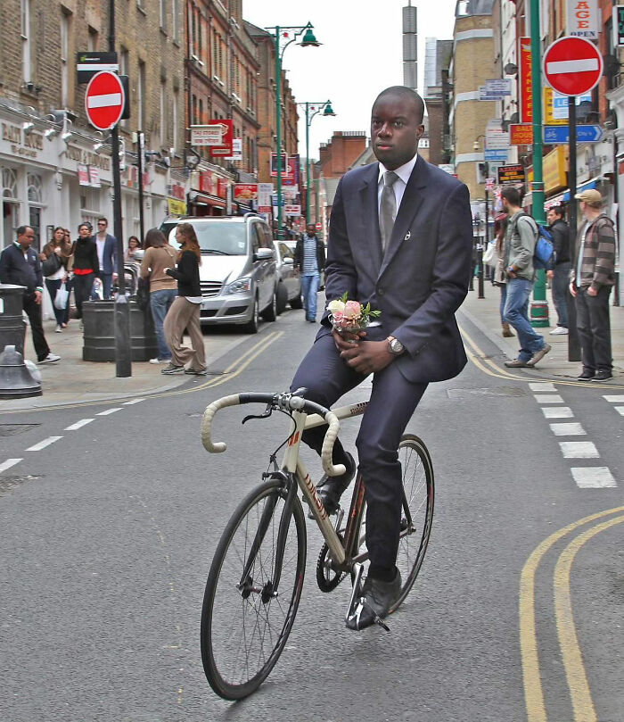 Man in a suit rides a bicycle holding flowers on a bustling street, showcasing pure street photography.