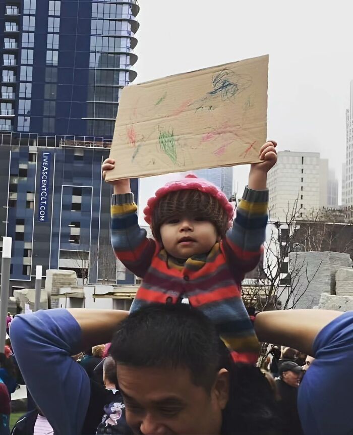 Child on shoulders holding a cardboard sign in a busy cityscape, exemplifying captivating street photography.