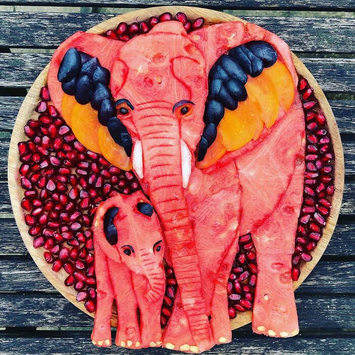 Healthy eating art featuring elephants carved from fruit, surrounded by pomegranate seeds on a wooden plate.