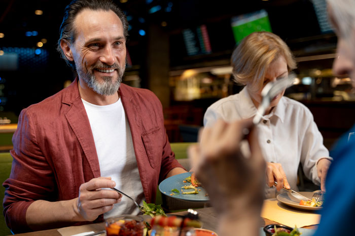 Man in red jacket smiling during brunch, with in-laws, in a warmly lit restaurant setting.