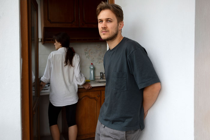 Man standing against a wall, looking away, while a woman is in the kitchen.