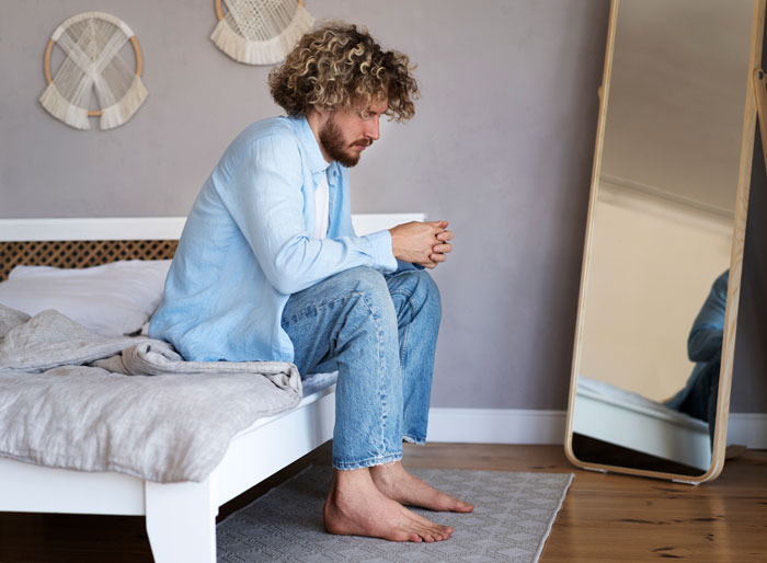 Man in casual clothes sitting on bed, looking thoughtful.