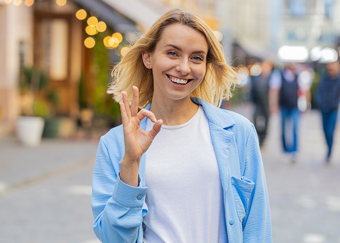 Smiling woman in a blue jacket making an OK gesture, embracing confidence and individuality in public.