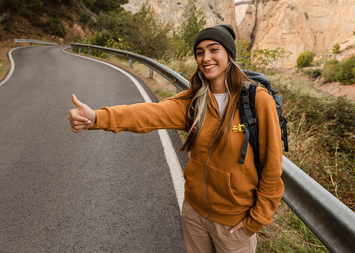 Woman enjoying outdoor adventure, smiling with a thumbs-up on a scenic road, showcasing confidence and self-love.