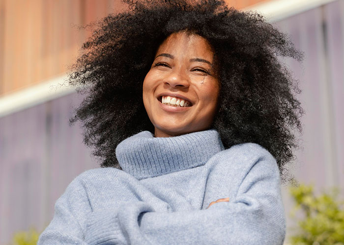 Woman confidently smiling, embracing her natural style with a cozy blue sweater outdoors.