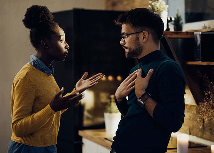 A woman in a yellow sweater discusses with a man in a kitchen setting, highlighting societal norms and self-acceptance.