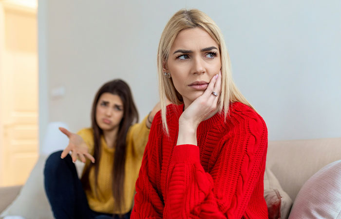 Woman in a red sweater looks upset, while another gestures in the background. Babysitter conflict depicted.