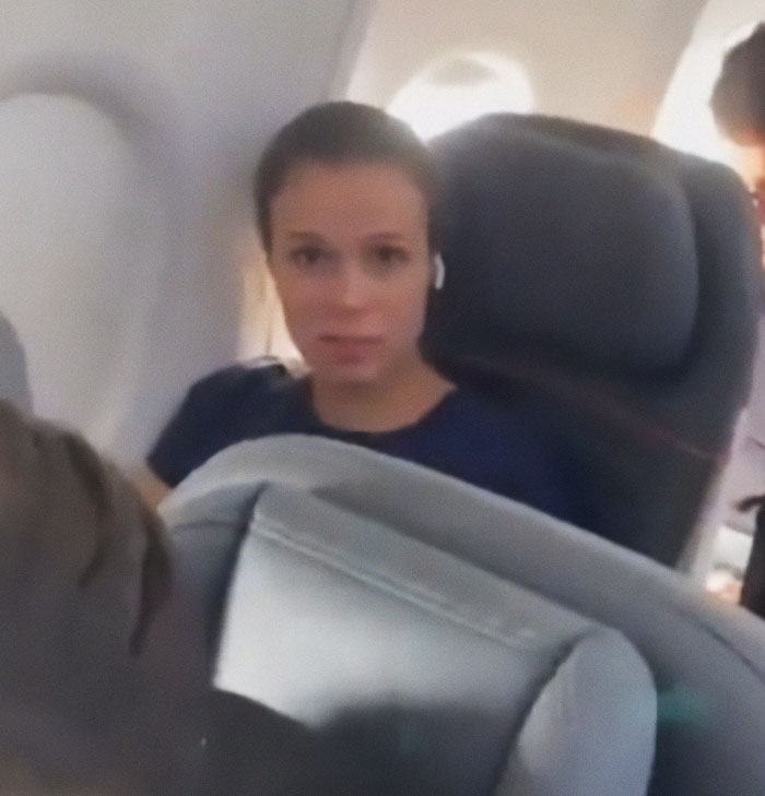 Woman in airplane seat, involved in seat swap dispute with crying child, wearing navy shirt and earbuds.