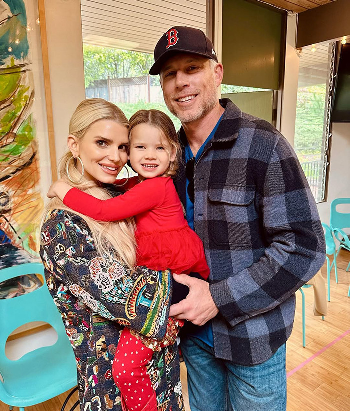 Family smiling together indoors, featuring Jessica Simpson and a man holding a young girl, both adults dressed casually.