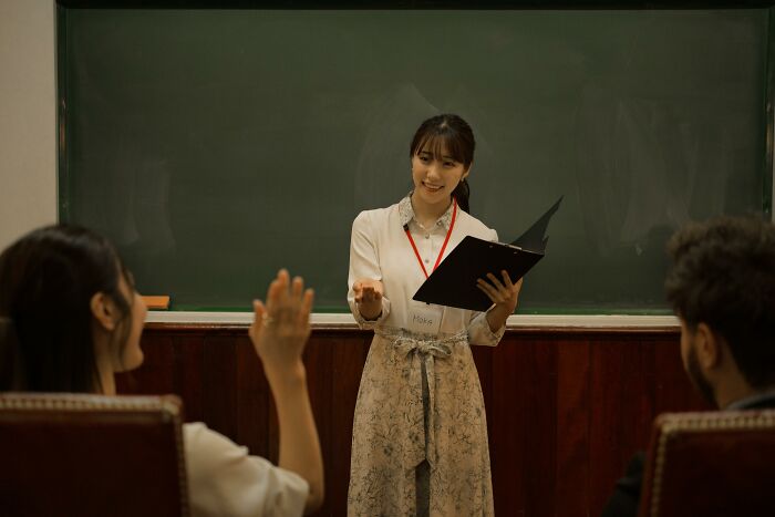 Woman holding a clipboard, speaking in front of a classroom, wearing a name tag labeled "Marie."