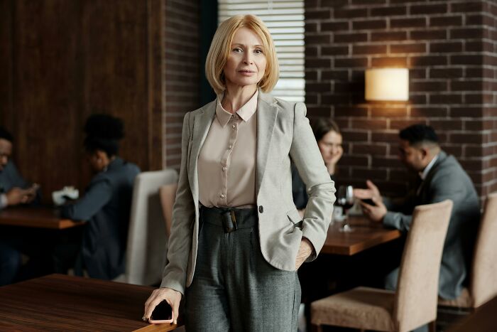 Woman in a cafe wearing a gray suit, holding a phone, with people in the background.