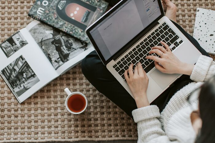 Person typing on a laptop at home, while sitting on a patterned carpet with a tea cup.