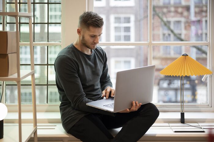 Man working on a laptop by a window with a yellow lamp nearby.