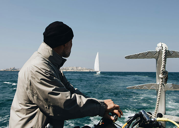 Man on a boat gazing at the sea, wearing a beanie and jacket, while a sailboat is visible in the distance.