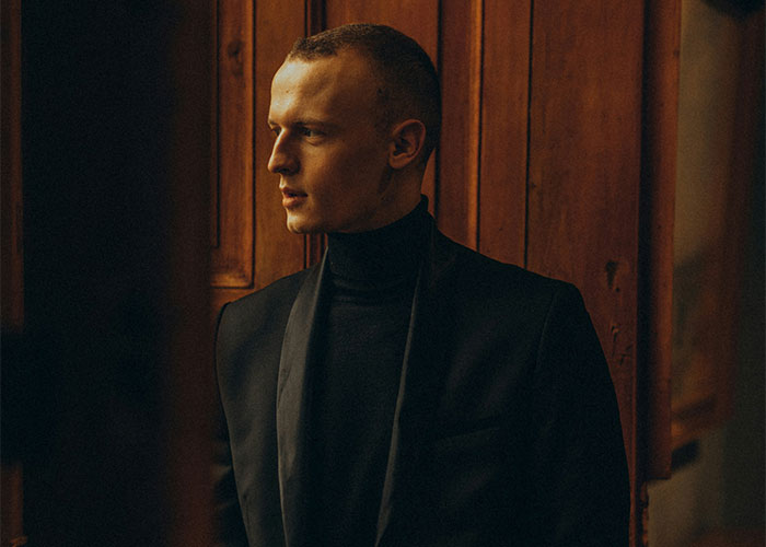 Man in a black suit in a dimly lit room, looking contemplative.