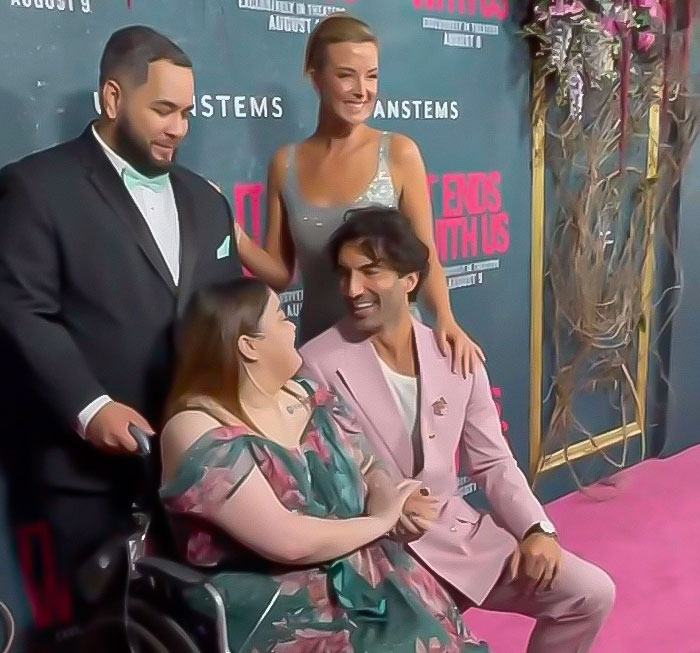 Group of people posing at a red carpet event, with a man in a pink suit smiling at a woman in a floral dress.