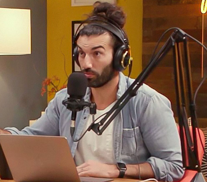 Man with headphones speaking into a microphone, seated at a desk with a laptop, in a casual studio setting.