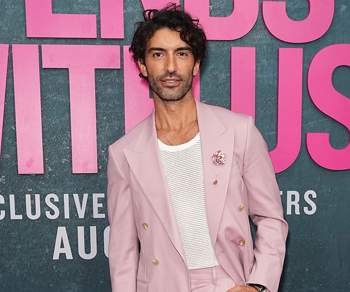 Justin Baldoni in a stylish pink suit at a public event, standing against a backdrop with bold text.