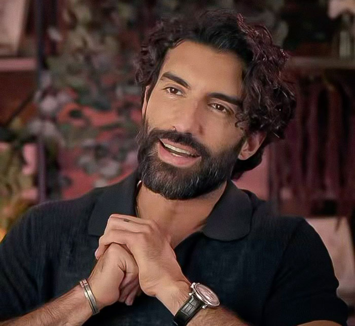 A person with a beard and curly hair, wearing a black shirt and watch, is sitting with hands clasped and smiling.