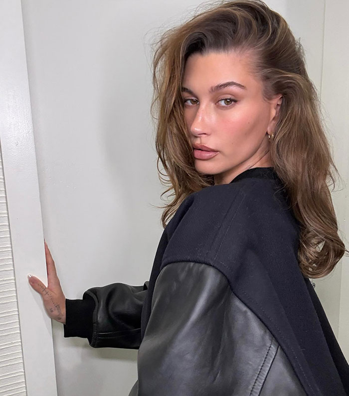 Hailey Bieber gazing back, wearing a black leather jacket, indoors.