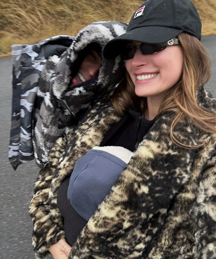 Hailey Bieber smiling in a fur coat next to a hooded person on a nature walk.