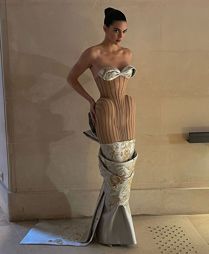 Kendall Jenner in a daring Paris Fashion Week dress with unique design elements against a plain background.