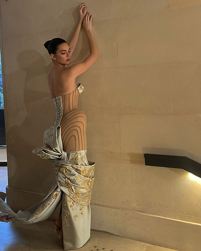 Kendall Jenner in a Paris Fashion Week dress with an intricate design, posing against a wall.