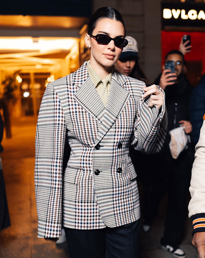 Kendall Jenner at Paris Fashion Week wearing a plaid suit and sunglasses, attracting attention from onlookers.