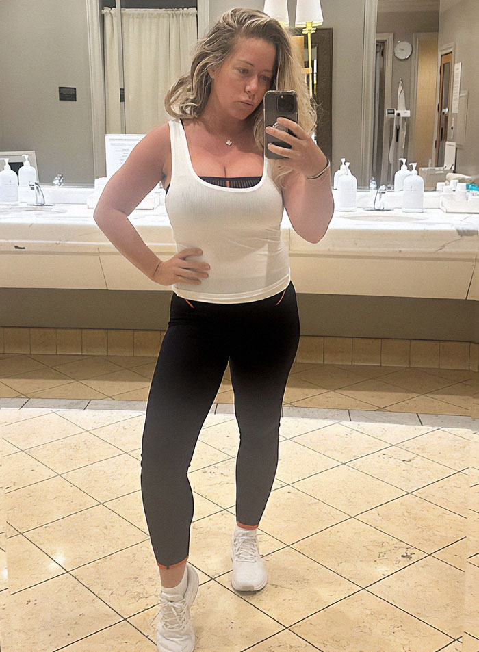 A woman takes a selfie in a bathroom wearing a white tank top and black leggings, addressing weight gain criticism.