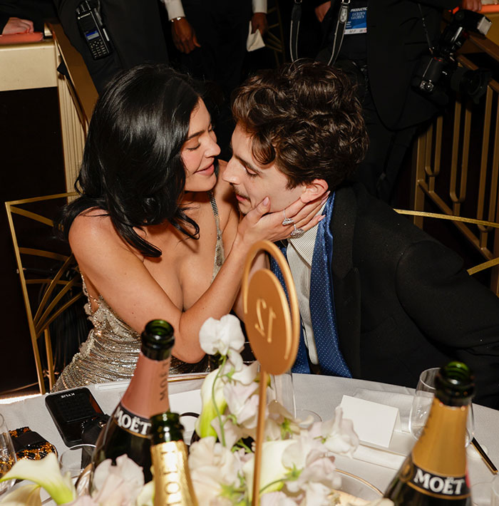 A woman in a sparkling dress sits closely with a man at a table, surrounded by flowers and champagne bottles, at an event.