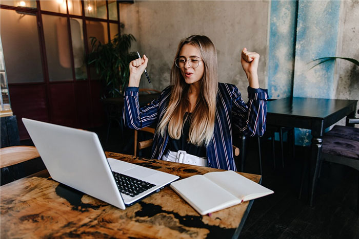 Young woman celebrating a success at her laptop, embodying wise observations and discoveries at a cafe table.