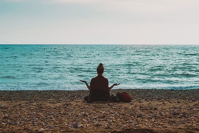 Person meditating on a pebble beach, facing the sea, embodying wise observations.