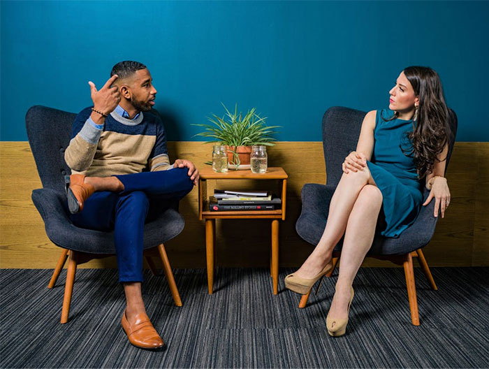 Two people engaged in conversation, sitting in chairs with a small table between them, sharing wise observations.