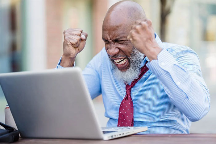 A man in a tie raises fists at a laptop, reacting to wise observations shared online.