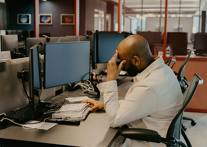 Person sitting at desk, appearing stressed, in an office setting, surrounded by computers, highlighting health hacks benefits.