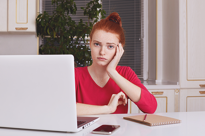 Woman in red shirt at desk with laptop, appearing thoughtful or concerned.