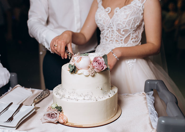 A bride and groom cutting a wedding cake, decorated with pink and white flowers on top.