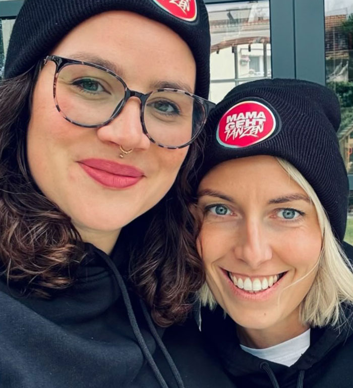 Two moms wearing matching beanies with the logo "Mama geht tanzen," smiling and redefining post-baby partying.