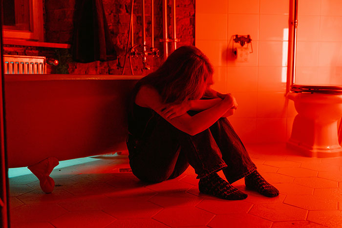 Woman sitting on bathroom floor, dimly lit in red, evokes a sense of tragedy and emotional struggle.