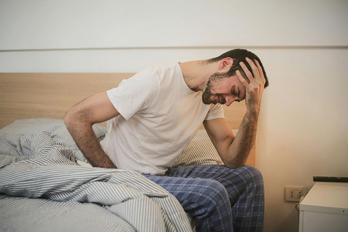 Man in distress sitting on bed, head in hand, wearing white shirt and plaid pants, reflecting on infidelity issues.