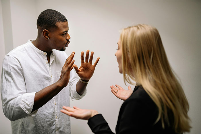 Man in a white shirt arguing with a woman in a black blazer, gesturing defensively in a heated discussion.