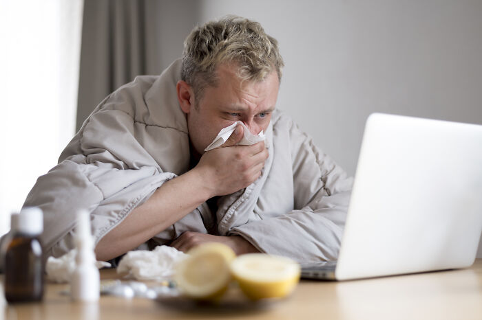 A person wrapped in a blanket, appearing unwell, sitting at a table with a laptop and cold remedies, highlighting middle-class challenges.