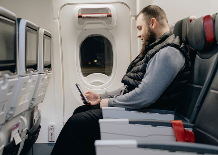 Man in window seat on airplane, wearing gray sweater and black vest, using his phone.