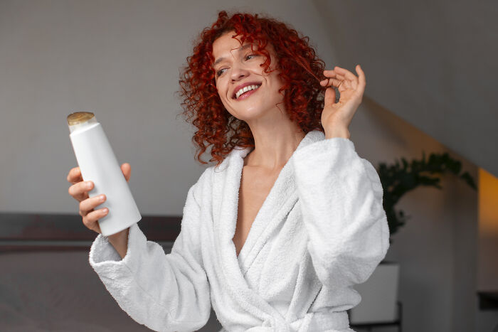 Person with curly hair in a white robe, smiling and holding an expensive shampoo bottle, in a cozy setting.