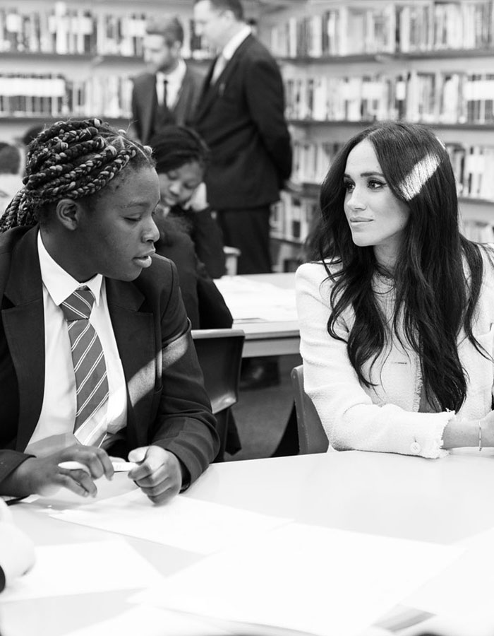 A woman converses with a student in a library, wearing formal attire, hinting at discussions involving workplace behavior.